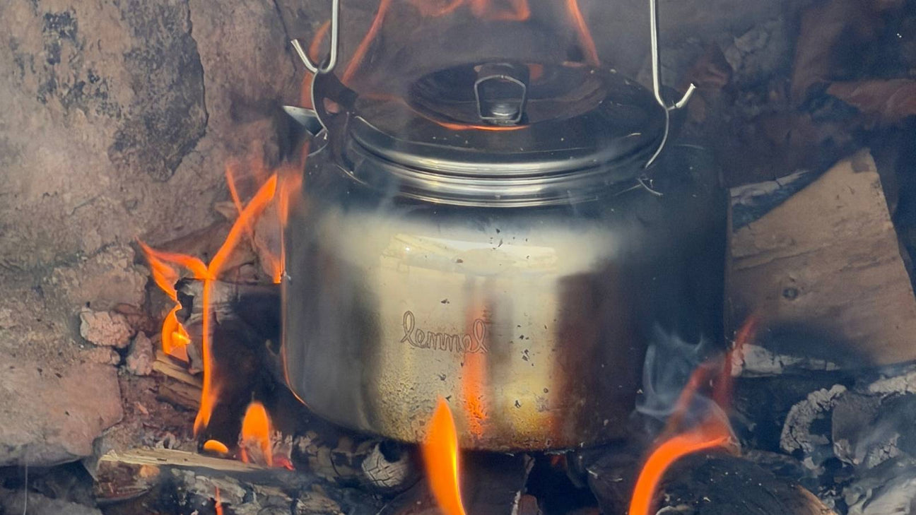 En rykande kaffepanna i rostfritt stål över en sprakande lägereld i skogen.