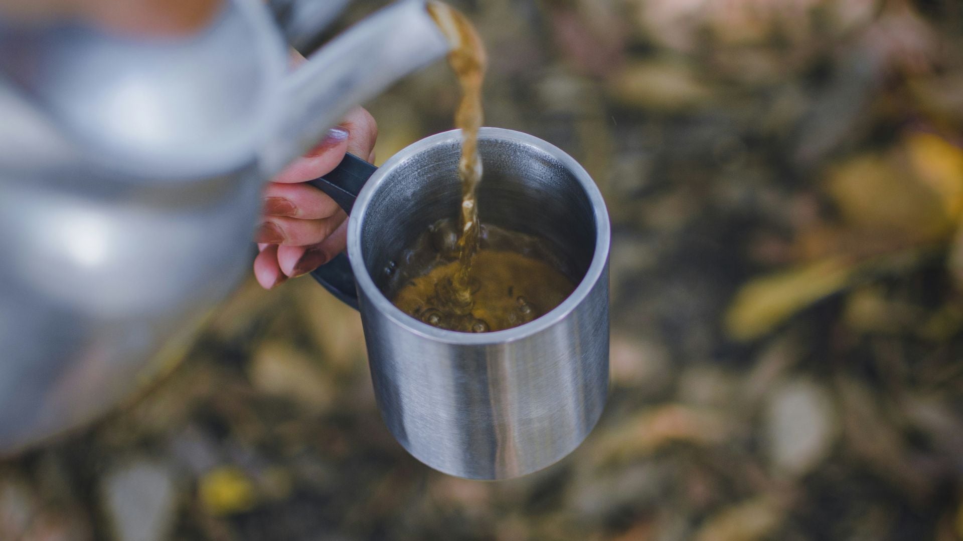 Närbild uppifrån där en person häller rykande hett kaffe från en klassisk kaffepanna i aluminium ner i en rostfri stålmugg med handtag, med en suddig bakgrund av skogsmark.
