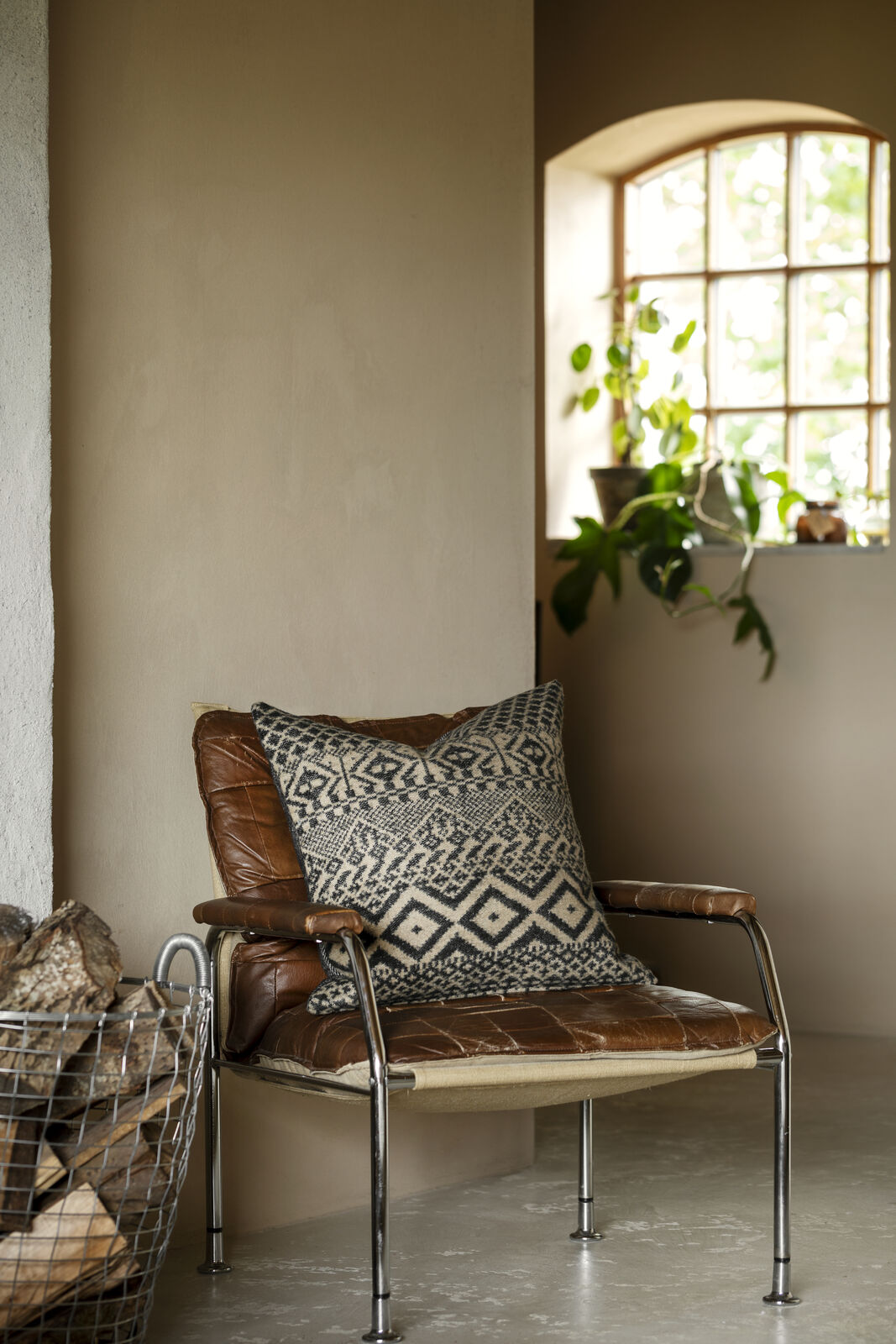 En brun läderfåtölj med en mönstrad kudde i ett rum med jordnära toner och en vedkorg i förgrunden.