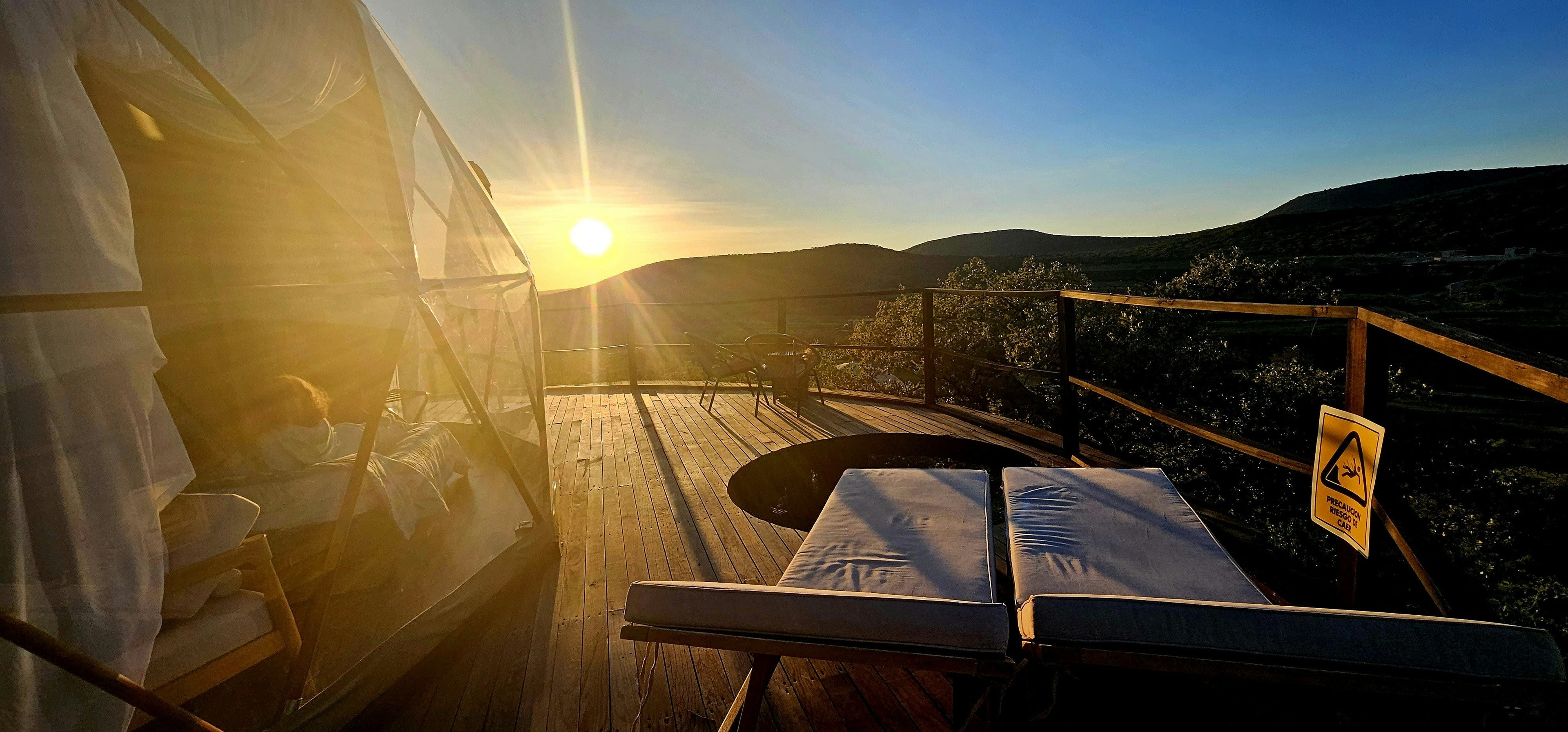 Ett genomskinligt glamping-tält på ett trädäck med solstolar och en inbyggd pool, med vacker utsikt över kullar i solnedgången.