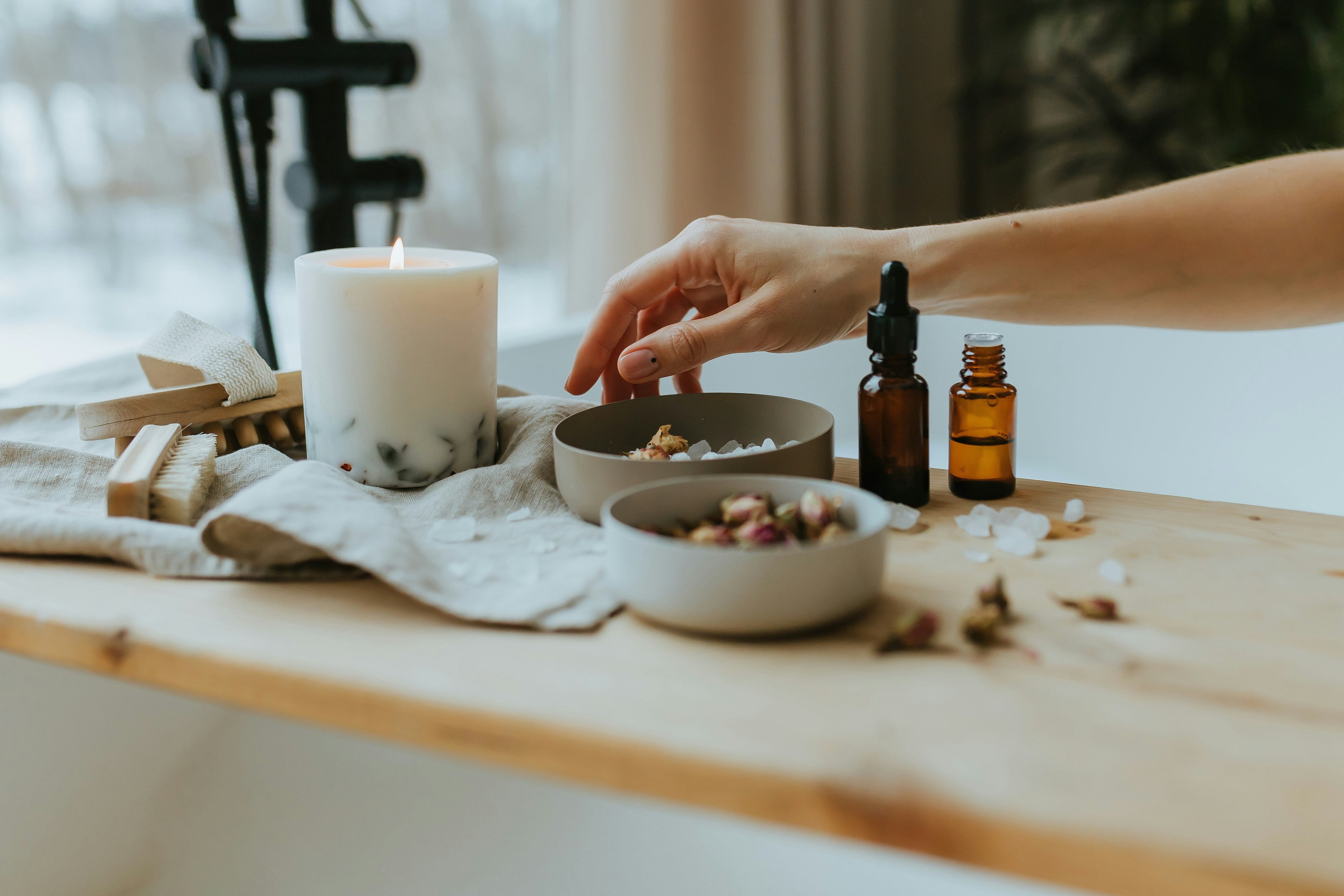 En hand som sträcker sig efter badsalt på en badkarsbricka i trä, tillsammans med ett tänt ljus och eteriska oljor.