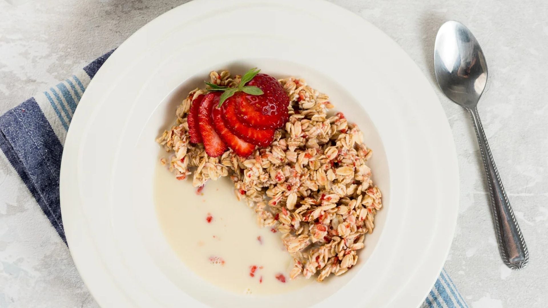 En skål med mûsli toppad med skivade färska jordgubbar, serverad med mjölk på en tallrik med en sked vid sidan.