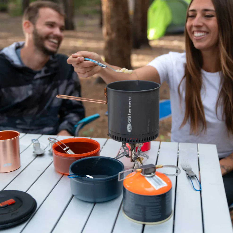 En man och en kvinna sitter vid ett campingbord i skogen och lagar mat med ett stormkök. Fokus ligger på en kastrull och färgglada skålar på bordet medan kvinnan smakar av maten med en sked.
