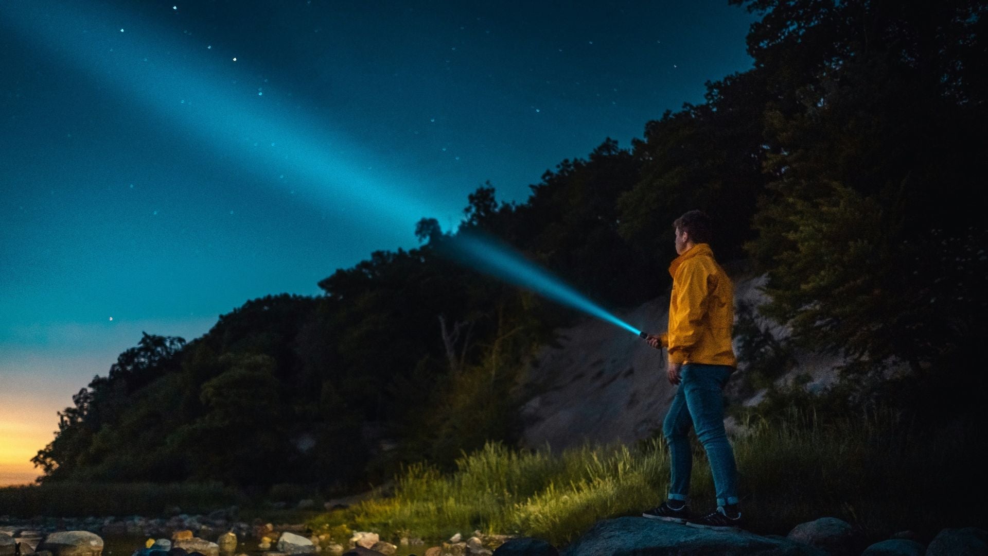 En person i gul jacka står vid en mörk skogsbryn på natten och riktar en kraftfull ficklampa med en blåaktig ljusstråle upp mot en stjärnklar himmel.
