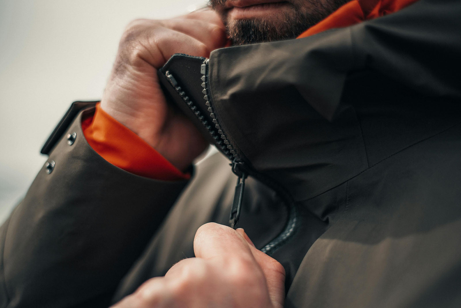 En närbild på en person som drar upp dragkedjan på en rejäl friluftshuva. Jackan är mörkgrön med ett kontrasterande orange foder som syns vid halsen och ärmsluten.