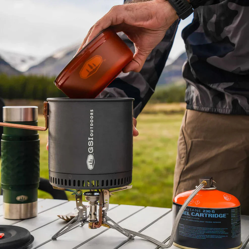 Närbild på matlagning utomhus med ett gaskök från GSI Outdoors placerat på ett fällbord. En person häller innehåll från en mindre orange kastrull ner i en större mörkgrå kastrull, med en orange gasbehållare och en grön termos i bakgrunden.