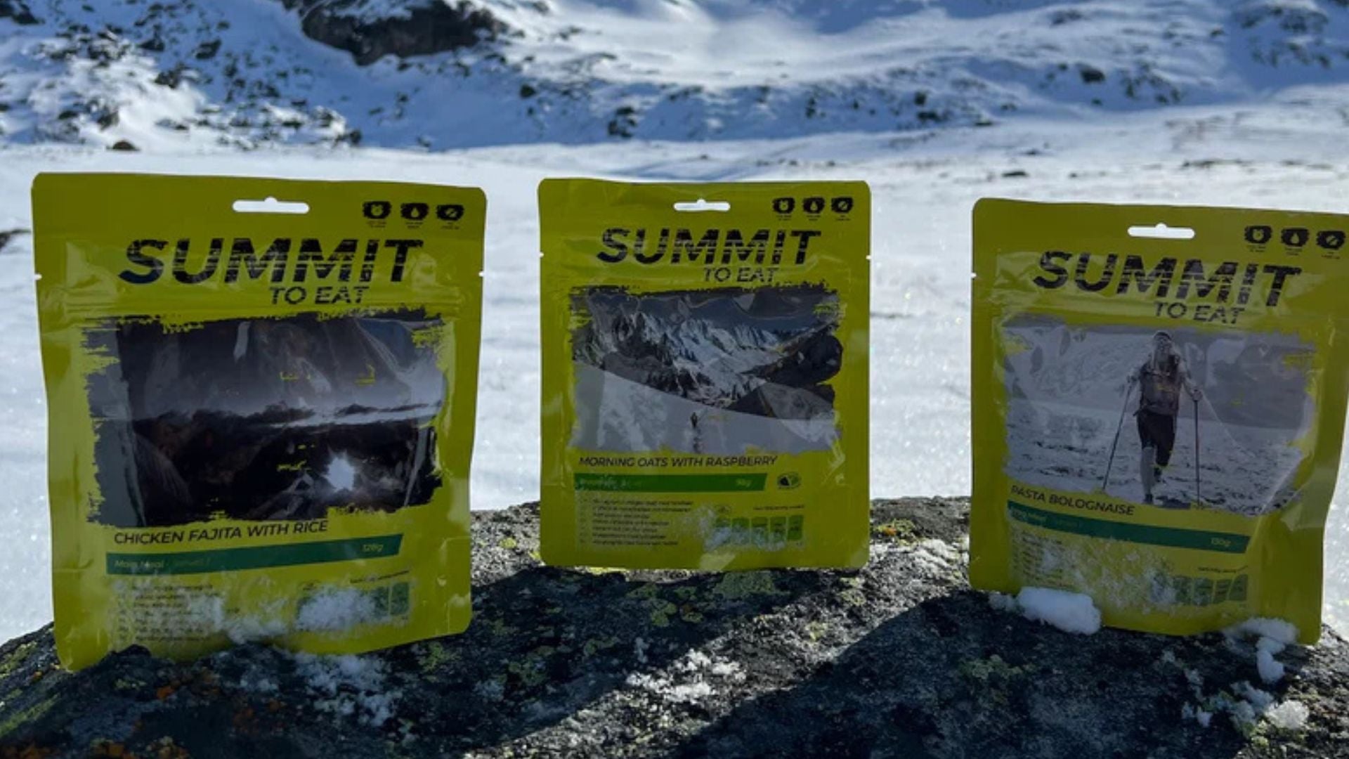 Tre gula påsar med frystorkad friluftsmat från Summit to Eat uppställda på en sten i ett vackert, snöigt fjällandskap.