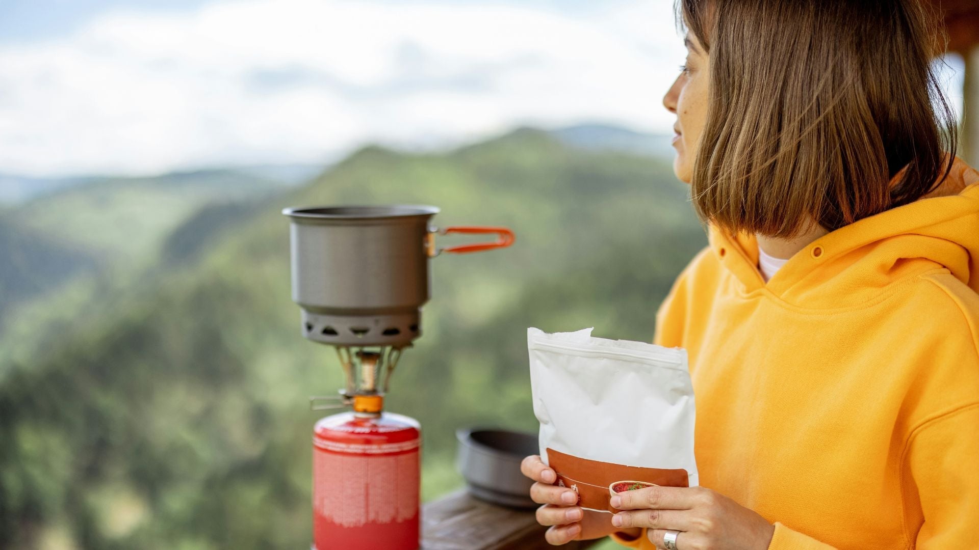 Logotypen för Adventure Menu över en naturbild där en person i gul luvtröja står vid ett stormkök och håller en påse med frystorkad mat, med utsikt över ett grönskande bergslandskap.