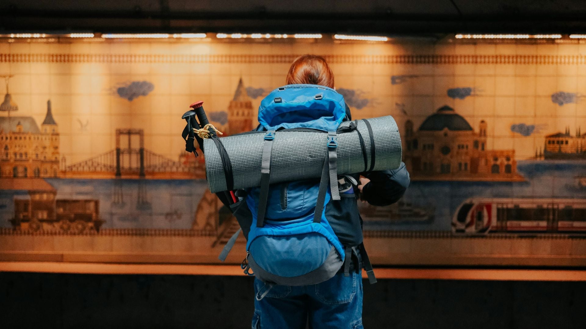 En person fotograferad bakifrån med en stor blå vandringsryggsäck, vandringsstavar och ett hoprullat liggunderlag, stående på en perrong framför en kaklad vägg med stadsmotiv.