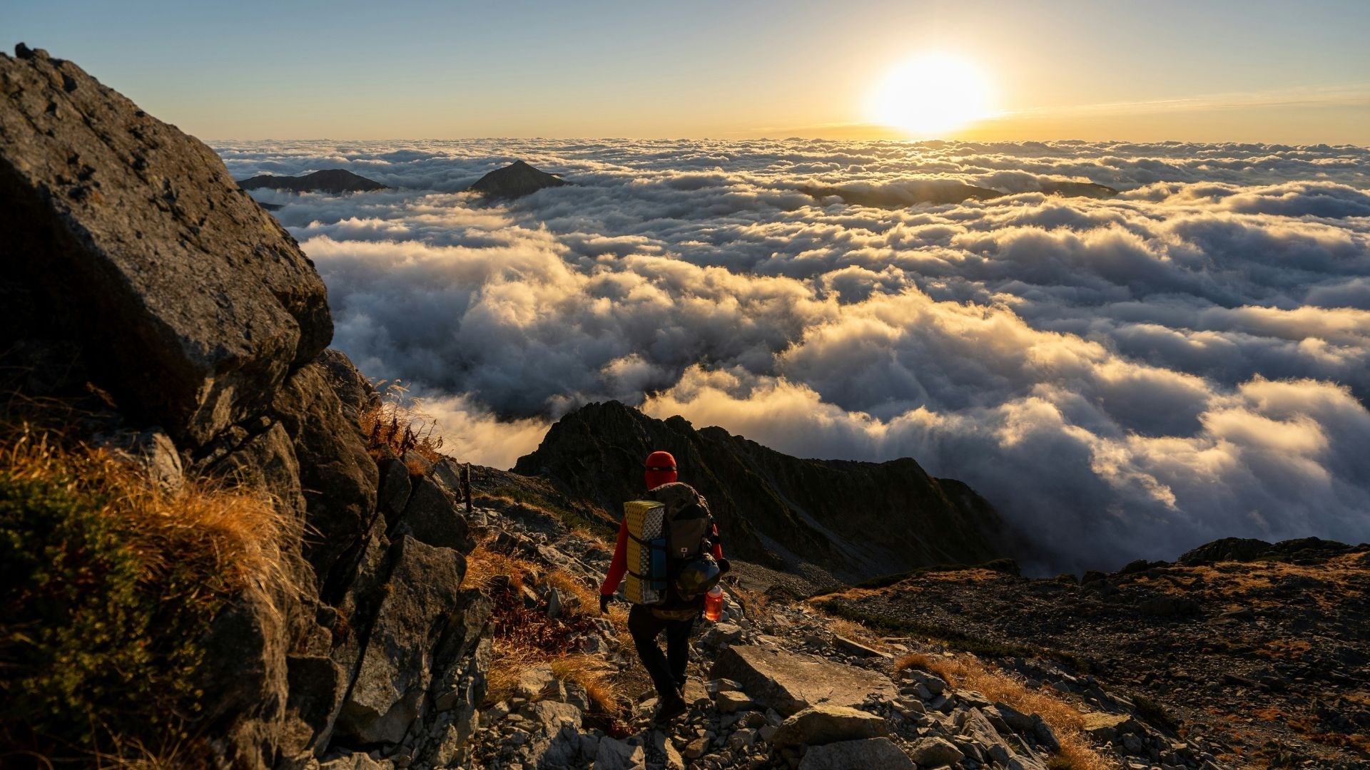 En vandrare med stor ryggsäck navigerar nerför en stenig bergsstig högt över ett utbrett molnhav i ett gyllene solljus. I det nedre högra hörnet syns logotypen för varumärket VAUDE.