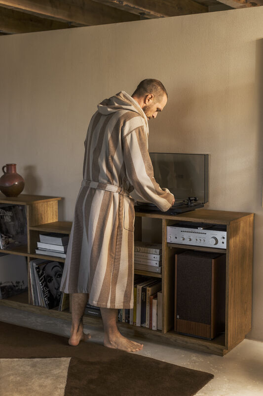 En man sedd från sidan står barfota i ett vardagsrum och justerar en vinylspelare på en träbänk. Han bär en knälång, randig morgonrock i beige och brunt från Gustav Ovland. Rummet har en varm, rustik känsla med synliga takbjälkar, en hylla fylld med vinylskivor och böcker, samt en stor mörk matta på golvet.