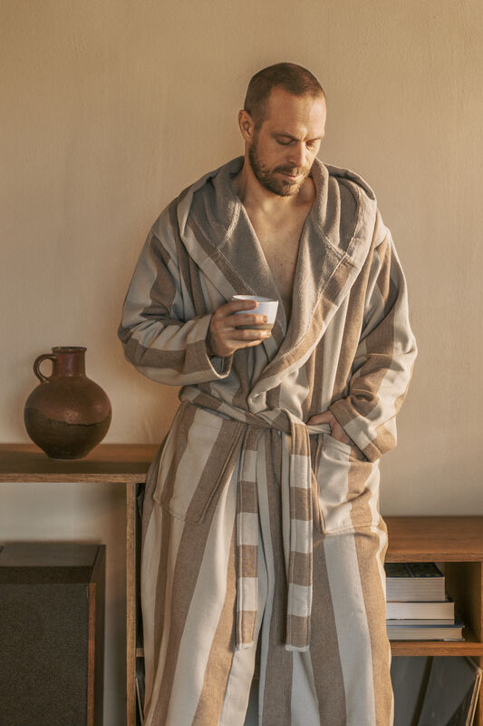 En man med skägg står lutad mot en trähylla klädd i en rymlig, randig morgonrock med huva från Gustav Ovland. Morgonrocken har breda vertikala ränder i beige och brunt. Han håller en keramisk kopp i handen och blicken är mjukt sänkt. Bredvid honom på hyllan står en brun keramikvas och några böcker, vilket skapar en harmonisk och avkopplande hemmiljö.