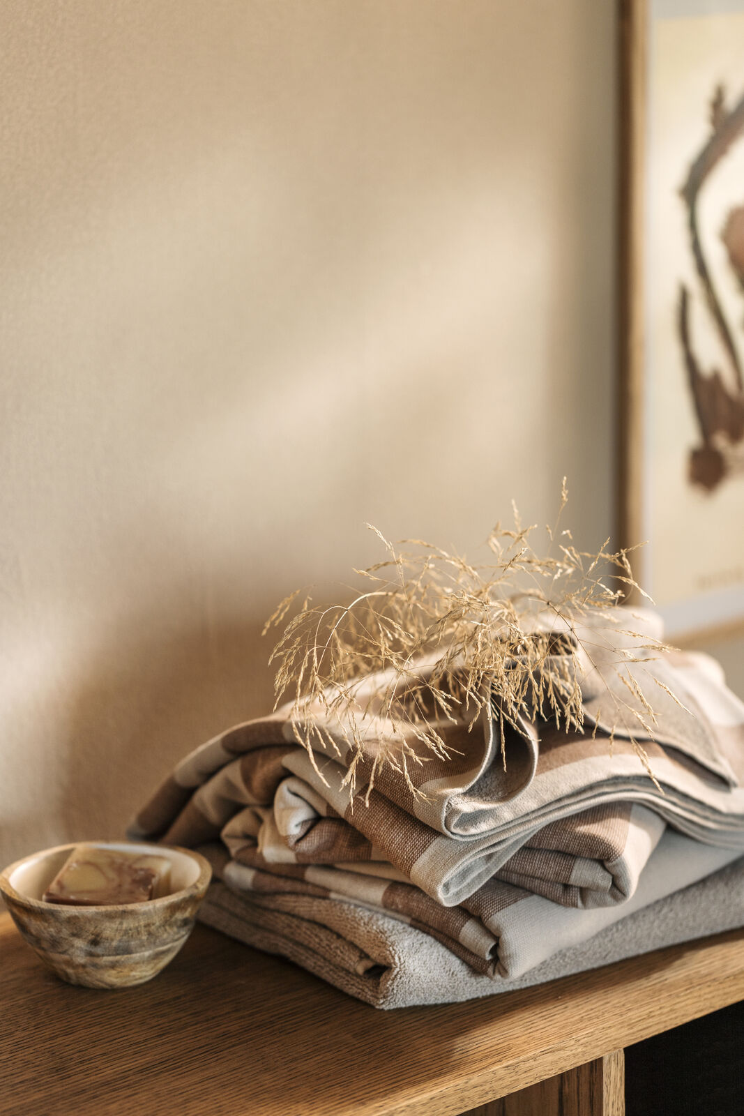 En mjukt vikt, randig handduk i jordnära bruna och beige toner från Gustav Ovland vilar på en bänk i trä. Ovanpå handduken ligger en torkad gräsvippa, och bredvid står en liten träskål med en bit tvål. Bakgrunden är varmt beige med mjukt solljus.