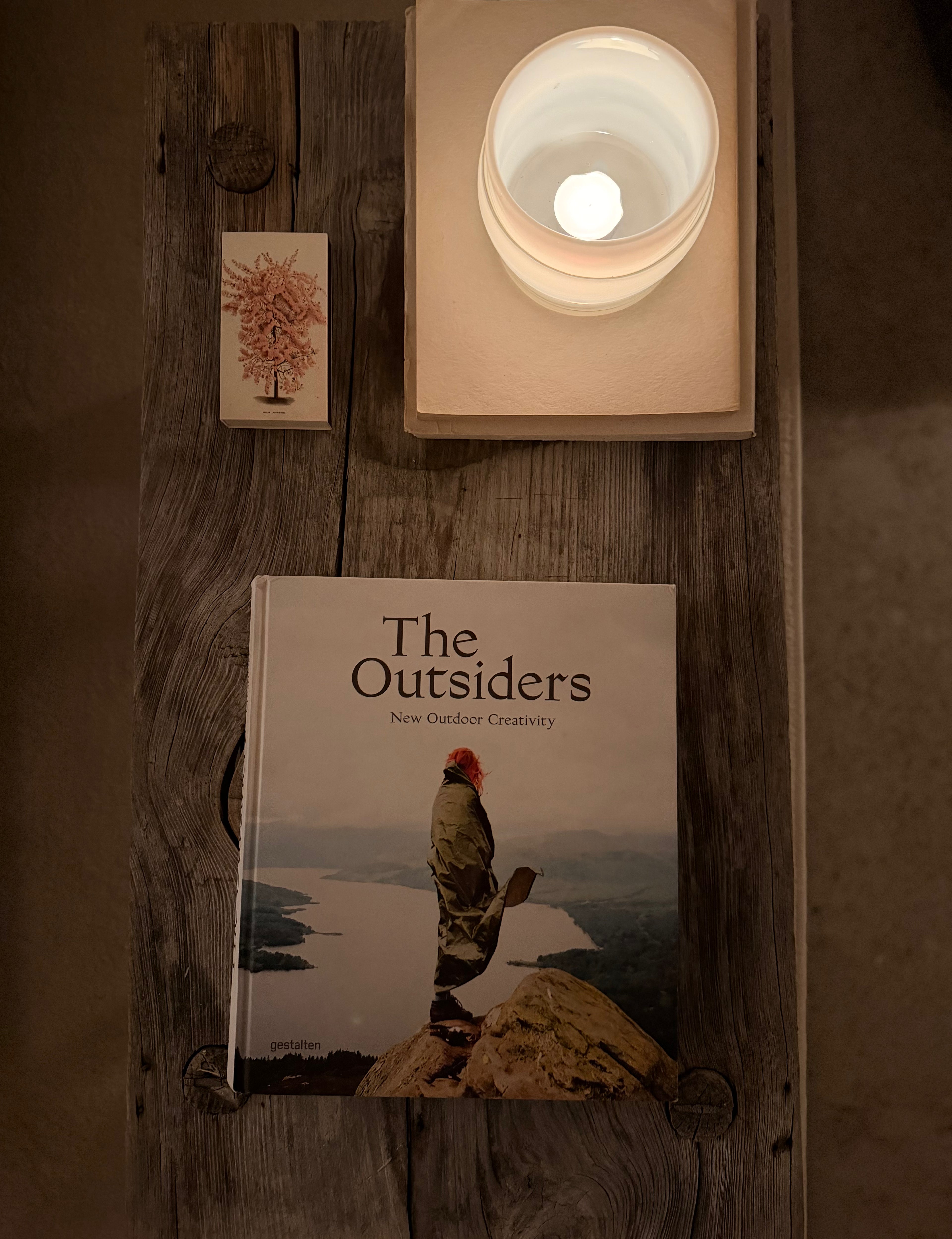 Boken 'The Outsiders: New Outdoor Creativity' placerad på ett rustikt träbord bredvid ett tänt ljus och ett litet konstkort. Bilden förmedlar bokens roll som ett kurerat objekt för inspiration och reflektion kring alternativ livsstil och estetik.