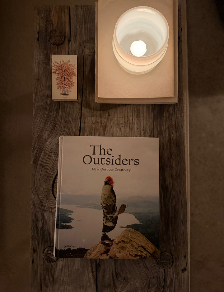 Boken 'The Outsiders: New Outdoor Creativity' placerad på ett rustikt träbord bredvid ett tänt ljus och ett litet konstkort. Bilden förmedlar bokens roll som ett kurerat objekt för inspiration och reflektion kring alternativ livsstil och estetik.