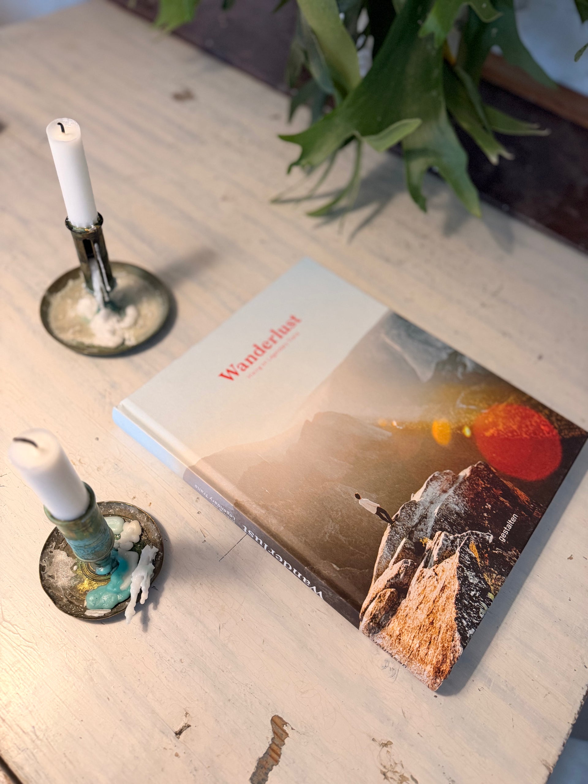 En inbunden bok med titeln ”Wanderlust” ligger på en vitmålad träyta bredvid två antikbehandlade ljusstakar med vita ljus. Bokens omslag visar en person som blickar ut över ett dramatiskt bergslandskap vid solnedgången. I bakgrunden syns de gröna bladen från en växt, vilket skapar en ombonad och inspirerande känsla för läsning och äventyr.