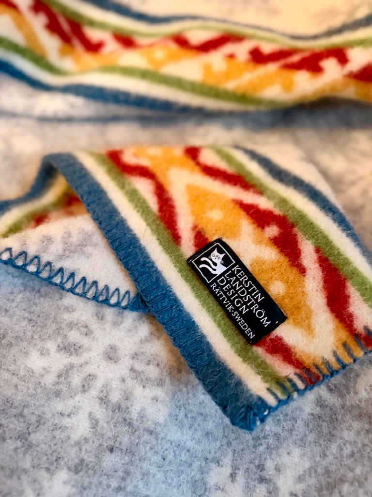 Närbild på en mönstrad yllefilt från Kerstin Landström Design. Filten har ett traditionellt dalainspirerat mönster i färgerna rött, gult, grönt och blått mot en vit botten. En svart tygetikett med vit text visar varumärkets namn och ”Rättvik - Sweden”. Filten ligger på ett grått underlag med ett diskret snöflingemönster.