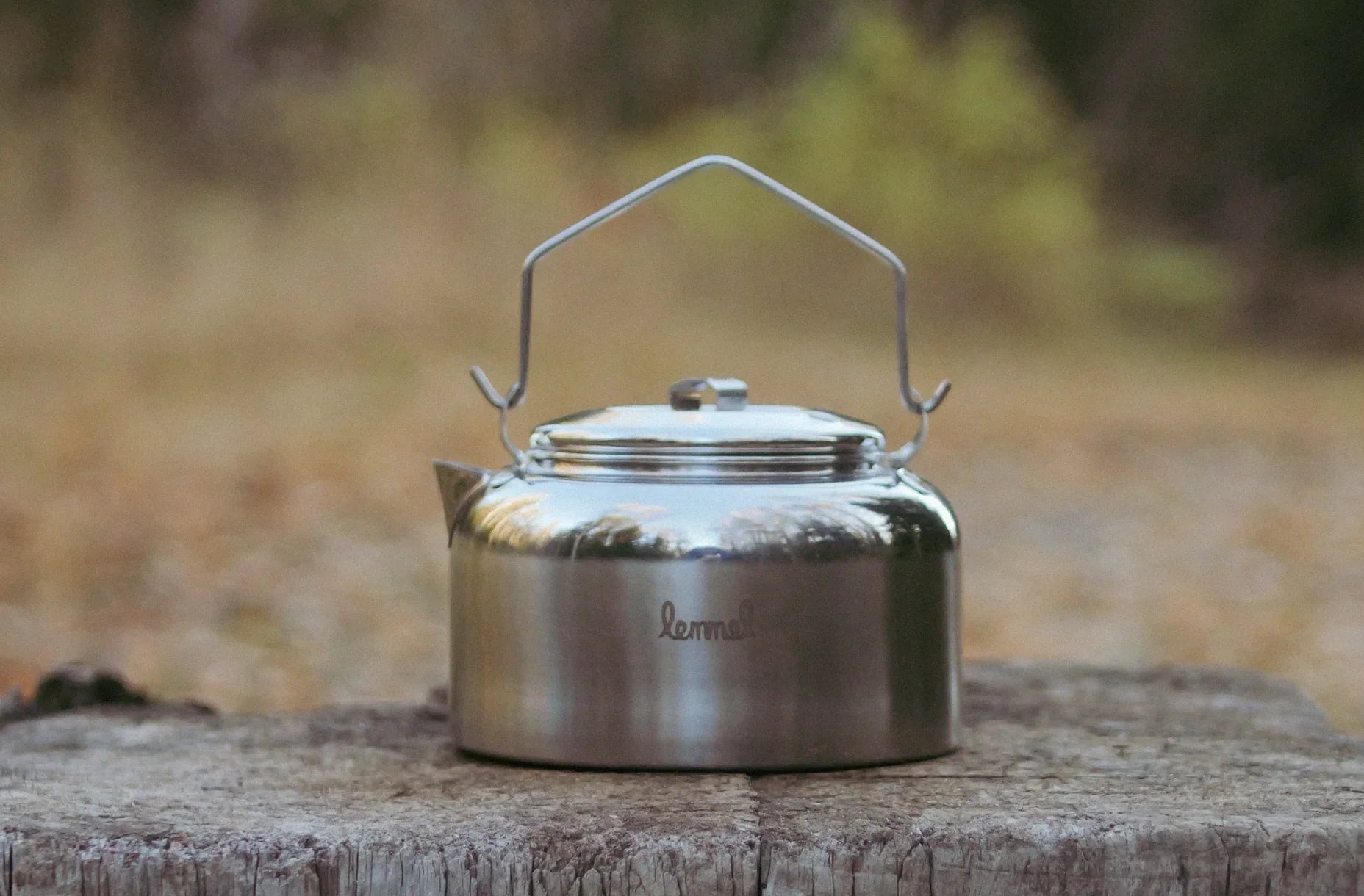 En klassisk kaffepanna i rostfritt stål med märket "Lemmel" ingraverat, placerad på en väderbiten trästubbe med en suddig skogsbakgrund i mjuka hösttoner.