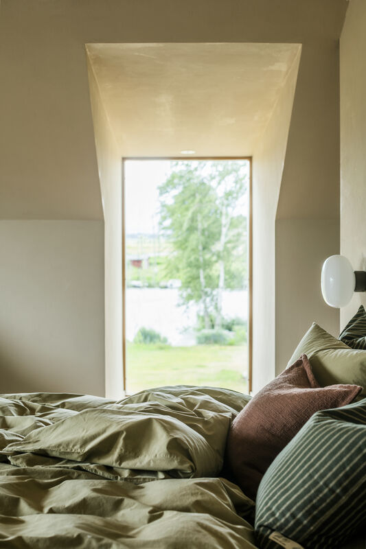 En harmonisk sovrumsmiljö med fokus på en bäddad säng i förgrunden. Sängen har mjuka sängkläder i dovt grönt, en brun prydnadskudde och en randig kudde i gröna nyanser. I bakgrunden finns ett djupt fönsterparti som ramar in en utsikt över en lummig trädgård med gröna träd och en skymt av vatten. Väggarna är i en varm, sandfärgad ton och en minimalistisk vit lampa är monterad på väggen bredvid sängen.