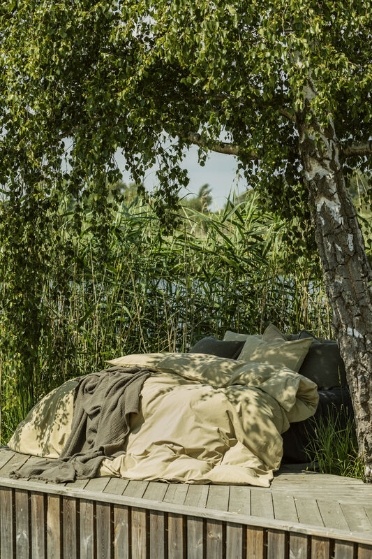 En bäddad säng med mjuka sängkläder i en dov grön nyans står placerad på ett trädäck utomhus. Sängen är placerad under en lummig björk vars grenar hänger ner som ett naturligt tak, och i bakgrunden syns högt grönt vass vid ett vattenbryn. Solljuset silas genom löven och skapar ett vackert mönster av skuggor över täcket.