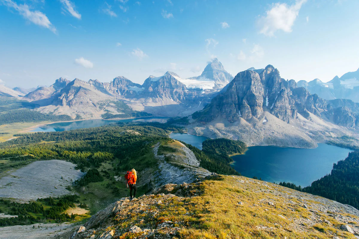En vandrare med röd ryggsäck står ensam på en bergskam och blickar ut över ett enormt alpint landskap med turkosa sjöar och spetsiga bergstoppar. En visuell skildring av outsiderns förmåga att kliva utanför systemet för att få en helhetsbild och upptäcka nya stigar i det outforskade.