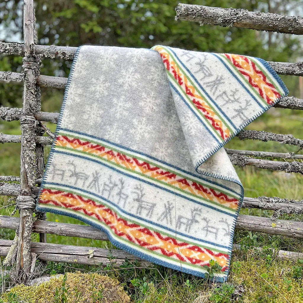 En mönstrad yllefilt hänger vikt över ett grått gammaldags gärdesgård i trä. Filten har en grå mittdel med vita snöflingor och breda bårder längs kanterna med renar och geometriska mönster i rött, gult, grönt och blått. Bakgrunden består av en lummig grön sommarhage.