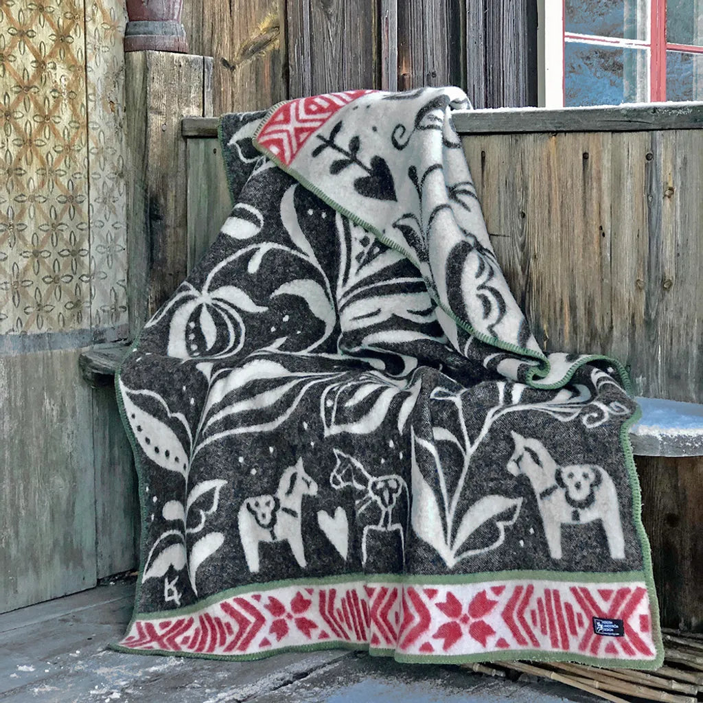 En mjukt vikt filt i jordnära bruna och beige ränder vilar på en bänk i trä. Ovanpå filten ligger en torkad gräsvippa som förstärker den naturnära känslan, och bredvid står en liten träskål med en bit tvål. Bakgrunden är en varmbeige vägg belyst av mjukt, naturligt ljus.